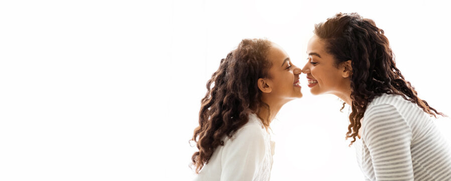 Loving african american mom and daughter touching each other with noses over white background, side view. Happy black young woman and school girl mother and child bonding over white
