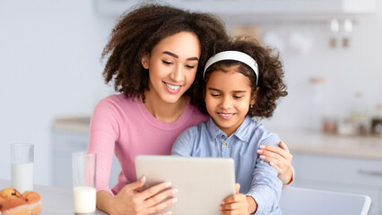 People And Technolody. Portrait of smiling African American little girl and young woman sitting at dinner table and using digital tablet, having breakfast, watching video or looking for new recipes