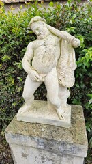 Herculaneum, Italy – 9 January 2025. A Roman marble statue in the House of the Deer shows Hercules resting with a lion skin draped over one arm, leaning slightly forward amid garden hedges.