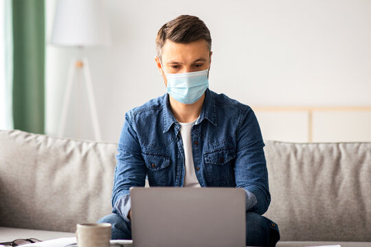 Businessman in protective face mask using laptop at home, sitting on couch in living room and sending his business partners and employees emails, copy space. Busniess and COVID-19 pandemic