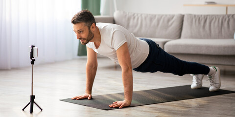 Motivated sporty middle-aged man training on fitness mat, broadcasting with smartphone, having online call with fitness trainer while staying home, copy space. Coach shooting video on mobile phone