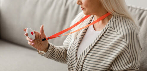 Emergency Button Concept. Portrait of serious senior lady holding personal alarm button, sitting on the at home alone. Older female looking at medical alert, selective focus
