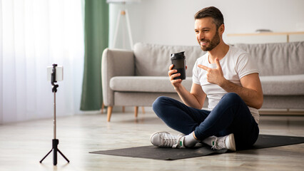 Cheerful middle-aged man fitness coach recommending protein drink for his followers or students while broadcasting from home, using mobile phone on tripod, pointing at bottle, copy space