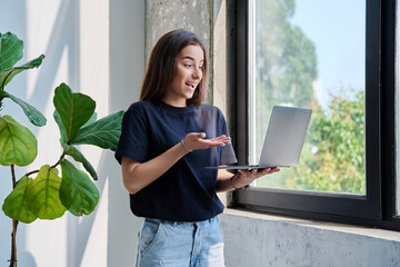 Young teenage girl using laptop for video chat conference standing at home