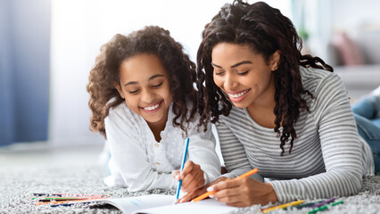 Smiling black mom and kid coloring book or painting with pencils together, laying on carpet in living room at home, closeup portrait. Art and craft family entertainment concept