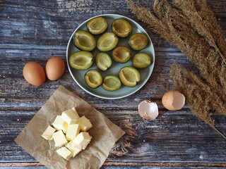 Fresh ingredients for homemade plum cake on rustic wooden table. Butter, eggs, and sliced plums prepared for baking.