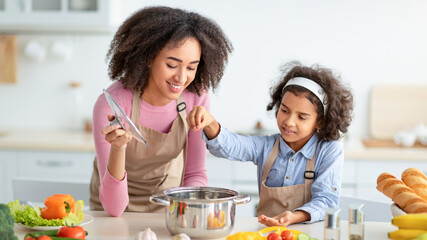 Bonding Concept. Cheerful african american mother helping her daughter to cook, teaching little girl how to prepare healthy vegetable soup, salting water in pot, cooking together at home.