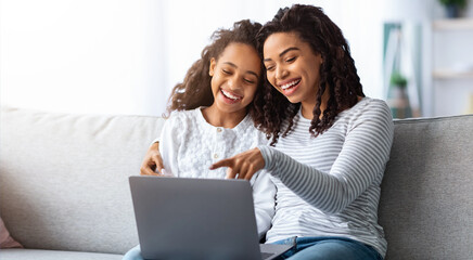 Fototapeta premium Excited african american mother and kid using laptop together while sitting on couch at home, closeup. Loving black mom and daughter playing online games on computer and hugging, copy space