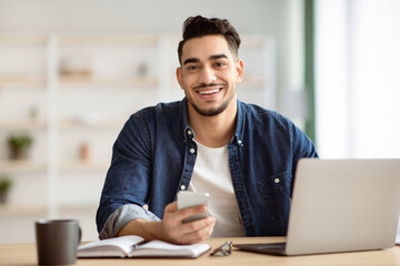 Obraz premium Smiling arab guy freelancer using smartphone while sitting at workdesk at home, working on laptop, copy space. Cheerful handsome middle-eastern man holding mobile phone, office interior