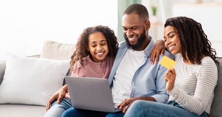 Easy Payment And Cashback Concept. Portrait of smiling African American family doing online shopping at home using laptop, woman holding credit card in hand, people sitting on couch at home