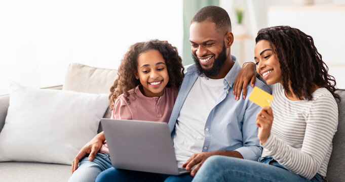 Easy Payment And Cashback Concept. Portrait of smiling African American family doing online shopping at home using laptop, woman holding credit card in hand, people sitting on couch at home