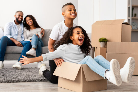 At New Apartment. Happy African American family having fun and celebrating moving day, excited brother riding his sister in cardboard box, parents hugging while sitting on the couch in background - Powered by Adobe
