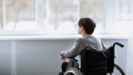Back view of disabled teenager in wheelchair looking out window, having depression at home, copy space. Paraplegic teen boy feeling stressed and upset, suffering from loneliness indoors © Prostock-studio