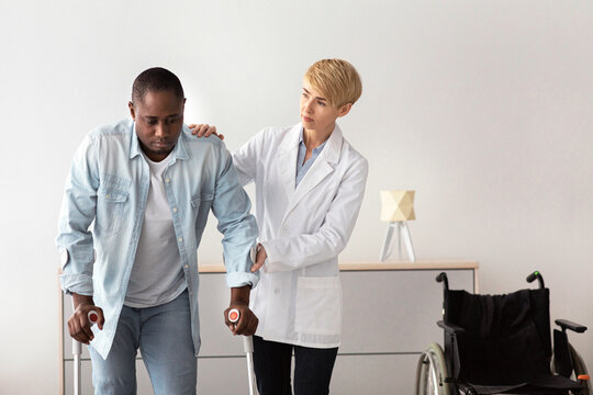 Recovery physiotherapy, medical service, specialist assistance in rehabilitation clinic. Serious adult female doctor help to sad middle aged african american man, to walk with crutches at home - Powered by Adobe