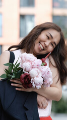 Fototapeta premium Portrait of a beautiful woman with a bouquet of peonies in her hands. Brunette in light summer clothes walks along a city street with a bouquet of flowers