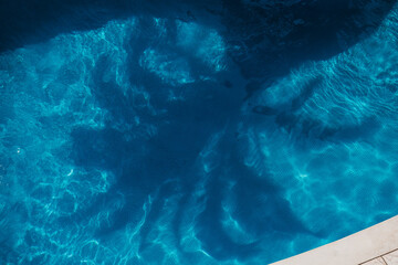 Clear blue water in a swimming pool and reflection of palm tree. The surface reflects sunlight, creating ripples and patterns. The pool edge is visible, made of light-colored tiles.