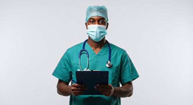 A doctor in scrubs and mask holding a clipboard looks at the camera on a white background