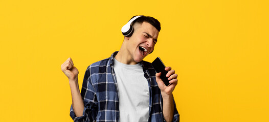 Emotional young man in headphones listening to music, singing song, using smartphone as mic on...