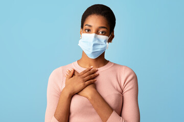 Young Blask Woman In Protective Medical Face Mask Holding Hands On Chest, African American Lady Grateful For Coronavirus Treatment And Vaccination, Standing Isolated On Blue Background, Copy Space © Prostock-studio
