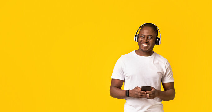 Positive emotions from favorite music at break. Happy middle aged african american male in white t-shirt and headphones and holds smartphone with app, isolated on orange background, studio shot