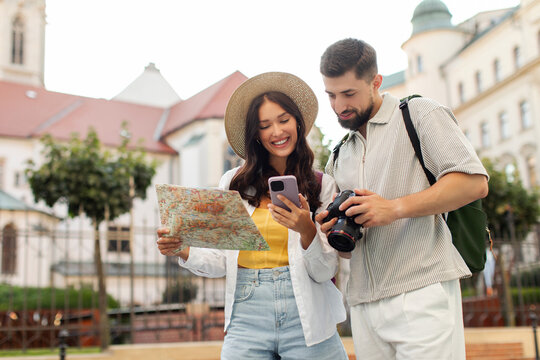 Couple of tourists consulting paper map and gps in smartphone online, standing in the street of an old town, sightseeing and enjoying trip
