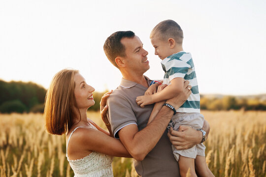 A joyful family spends time outdoors in a golden field during late afternoon. The father holds his smiling son, while the mother looks at them with affection.
