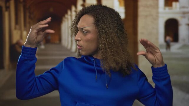 Woman making animated conversational hand gesture on historic street in blue hoodie; expressive communication.