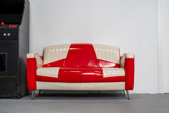 Retro red and white vinyl sofa with vintage diner style design placed indoors beside a classic arcade machine, symbolizing nostalgia, pop culture, leisure, retro interior decoration
