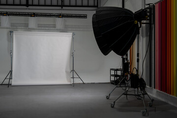 Professional photo studio setup with large softbox lighting, seamless white backdrop on stands, and colorful paper backgrounds stored on the side wall, ready for portrait photography, product shoots