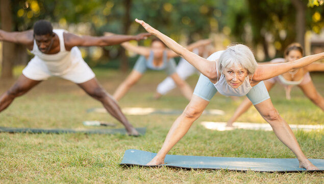 Multinational people of different ages in park practicing yoga. Amateurs performing gymnastic. Senior woman yoga master and followers do Prasarita Padottanasana. People practice during sport in nature - Powered by Adobe