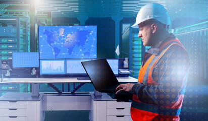 Engineer in helmet and safety vest using laptop in data center control room with multiple monitors showing world map and analytics, IT infrastructure, cybersecurity, networking.