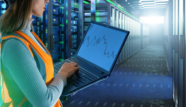 Woman in safety vest monitoring server racks with laptop showing data charts, managing IT infrastructure, cloud computing, cybersecurity, networking, data analysis.