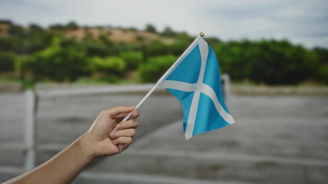 Man holding a scottish flag on an outdoor tennis court showing national pride in scotland with a blurred green background.