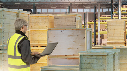Warehouse worker in safety vest using laptop for cargo management among wooden crates, concept of logistics, inventory, supply chain, storage. Logistics, Supply Chain, Inventory.
