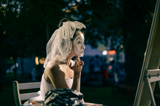actress in white wedding dress applies theatrical makeup to face sitting at mirror. portrait mime close-up side view