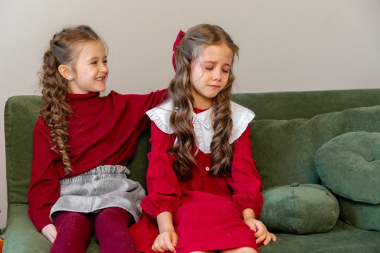 Two little girls sisters are sitting on a sofa near a Christmas tree. One girl is upset and cries, the other one hugs her and calms her down. Childhood and friendship relations concept.