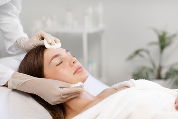 Young woman having facial beauty treatment, beautician cleaning and touching female face with cotton pads, free space