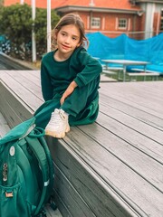 A schoolgirl in a green jacket sits on a wooden deck with her green backpack. The background shows the school building, reflecting an education concept,starting grade at a primary school in Australia