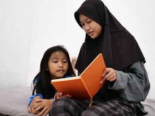 Asian muslim teenage girl reading book for her little sister before bedtime