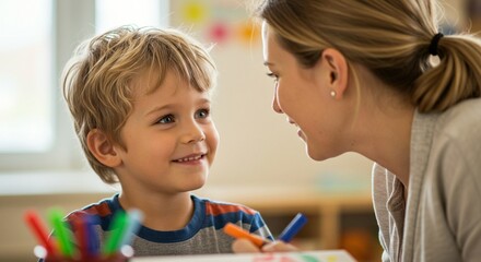 A smiling young boy with blonde hair looks up at a woman with a ponytail, while drawing with colorful markers at a table.