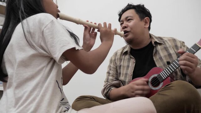 Asian man teaching his daughter how to play recorder flute while he plays ukulele. Music teacher and student