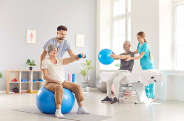 Obraz na płótnie Canvas Health care workers helping mature people to do sport exercises with dumbbells and fit ball in gym. Physiotherapist helping retired man and woman in rehab in rehabilitation center.