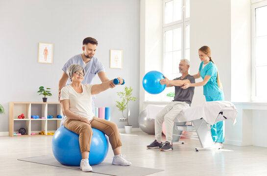 Health care workers helping mature people to do sport exercises with dumbbells and fit ball in gym. Physiotherapist helping retired man and woman in rehab in rehabilitation center. - Powered by Adobe