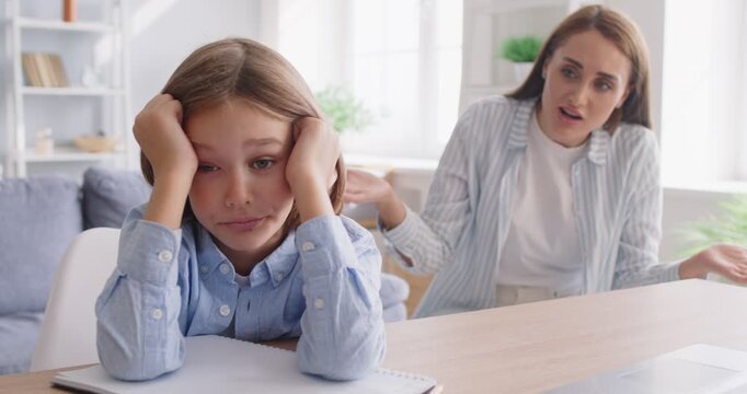 Frustrated apathetic indifferent long haired child kid boy sitting at table and listening to angry mad mother telling him off for misbehavior and bad grades. Family, education, school problems concept
