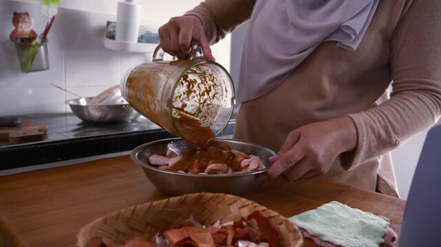 Asian muslim woman pouring herbs and spices seasoning from blender over chicken meat in bowl, marinating process