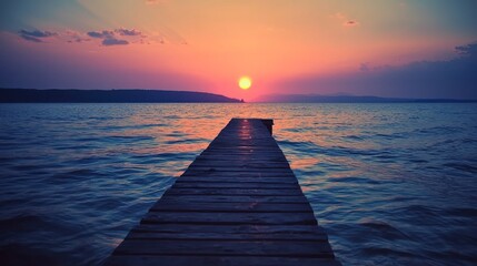Obraz premium A wooden pier extending into the calm, blue waters of a lake at sunset, with a vibrant orange and pink sky and a single, bright yellow sun in the distance.