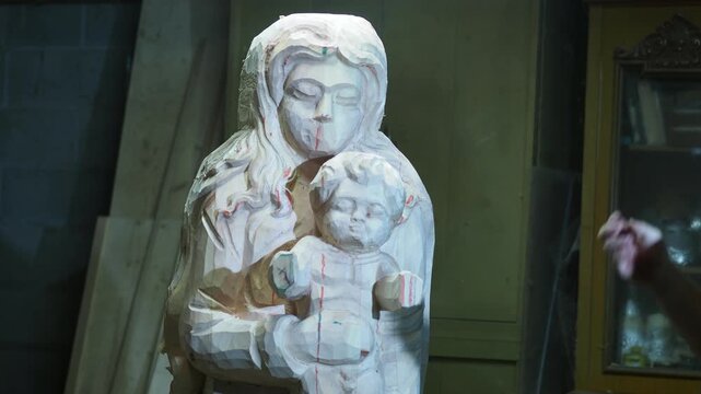 A partially carved wooden statue of the Virgin Mary holding the Child is shown in progress. The artisan uses hand tools to shape the figure, leaving visible chisel marks and guide lines.
