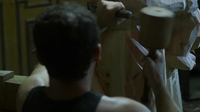 Craftsman strikes a chisel with a wooden mallet while carving details into a large wooden statue of the Virgin and Child, focusing on precision work inside a dimly lit workshop.