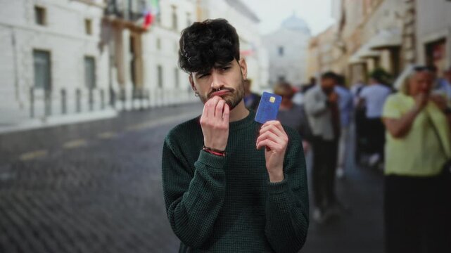 Young man holding creditcard on urban street pondering financial decision considering people and city backdrop suggesting outdoor public environment and thoughtful expression