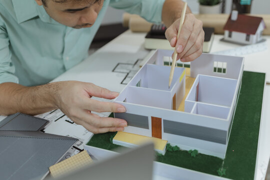 Asian male architect is analyzing a modern house model while holding the roof structure and pointing at layout with a pencil. Concept of architectural planning, property design, and home development.